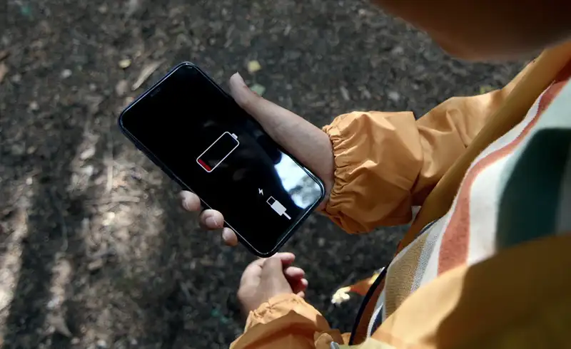 iPhone tinut in maini pe un drum forestier, ecranul afiseaza iconul de baterie aproape goala cu zona rosie si simbol de incarcare necesar — telefon care s-a oprit brusc desi mai avea procent in indicator
