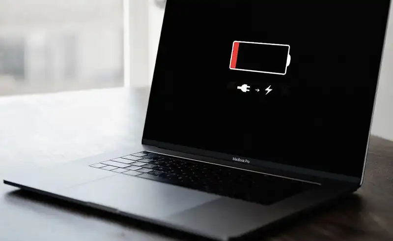 MacBook Pro deschis pe o masa intunecata cu ecranul aratand iconul de baterie aproape goala plus simbolul de alimentare necesara — Mac-ul s-a oprit brusc desi mai avea procent in indicator
