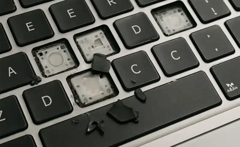 Macro pe o tastatură MacBook cu mai multe taste scoase și mecanismul scissor expus, chunks de plastic negru rupte împrăștiate pe suprafață — exemplu real de tastatură distrusă din cauza unui mecanism aftermarket inferior